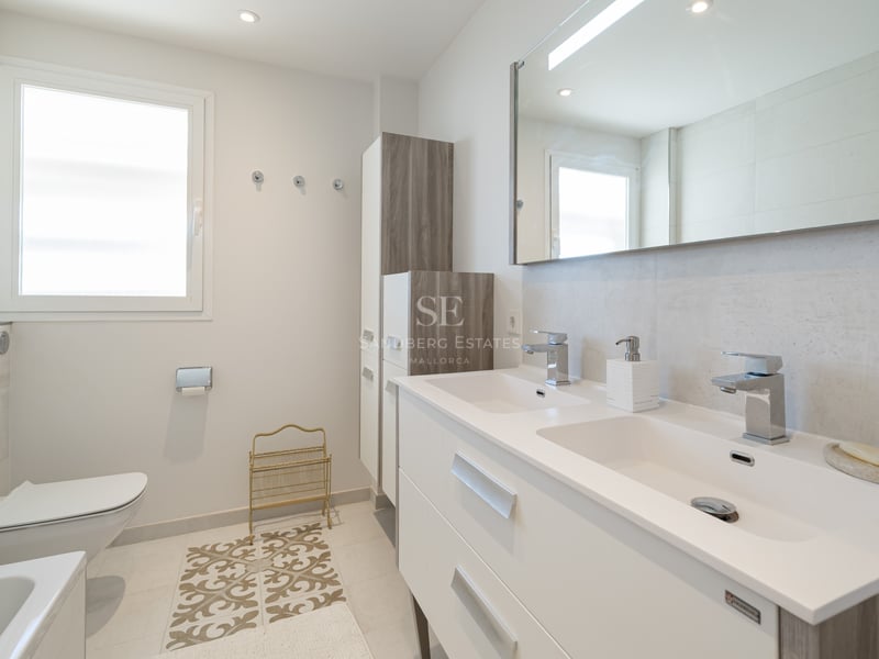 Salle de bain moderne lumineuse avec double vasque blanche, grand miroir, WC suspendu et lumière naturelle.