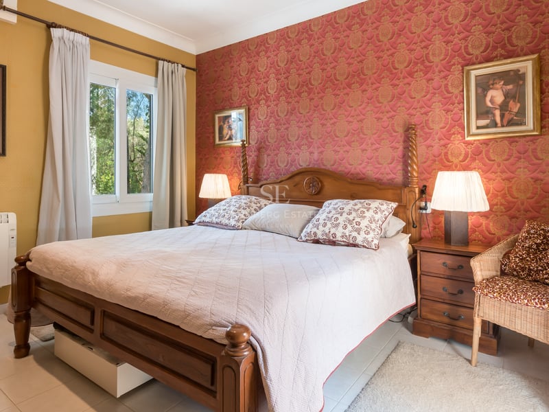 Chambre traditionnelle avec lit en bois, papier peint damas rouge et fenêtre donnant sur des arbres.