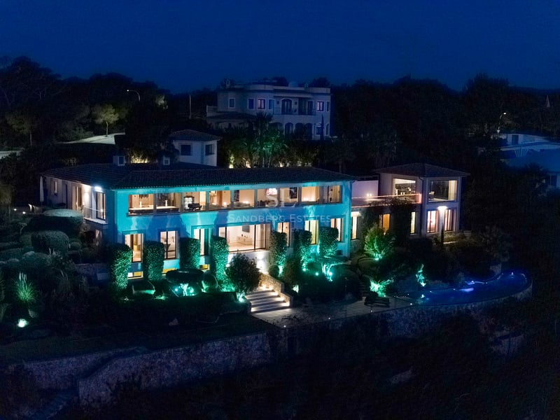 Vue nocturne d'une villa de luxe sur deux niveaux avec intérieurs éclairés, piscine et jardins paysagers.