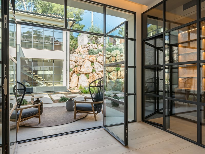 Vue intérieure vers une terrasse avec portes en acier noir, fauteuils design et mur en pierre naturelle.