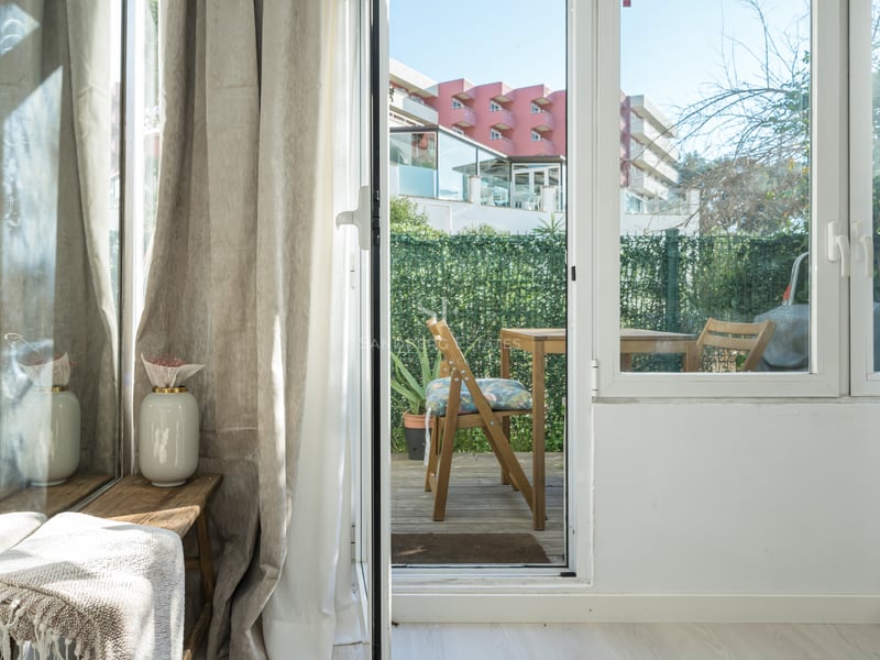 Vue à travers une porte vitrée sur une terrasse en bois avec mobilier d'extérieur et haie d'intimité.