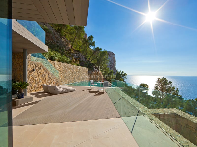 Terrasse de villa moderne avec piscine à débordement, deck en bois, garde-corps en verre et vue panoramique sur la mer.