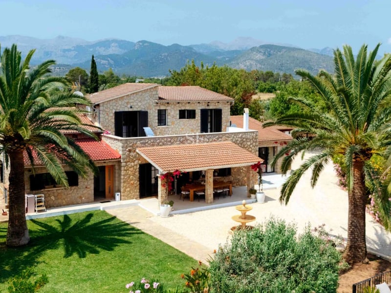 Villa traditionnelle en pierre avec palmiers, pelouse verdoyante et montagnes en arrière-plan.
