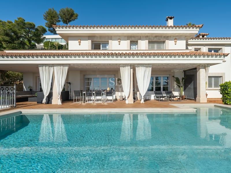 Vue d'une villa de luxe blanche avec grande piscine, terrasse couverte et rideaux blancs sous un ciel bleu.
