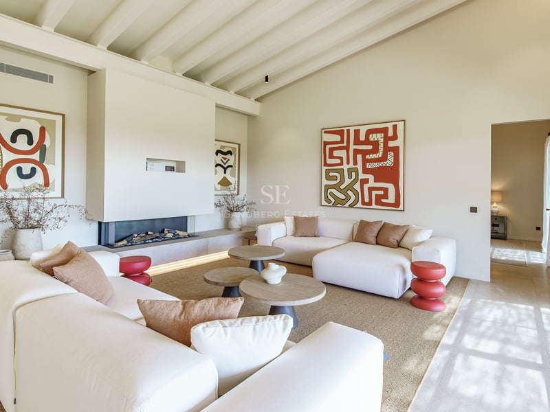 Salón luminoso con techos altos de vigas, chimenea moderna integrada, sofás blancos y arte abstracto en tonos tierra.