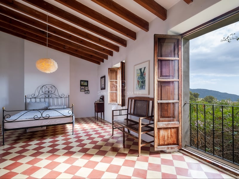 Chambre avec sol en damier, lit en fer forgé, poutres en bois et porte ouverte sur des montagnes verdoyantes.