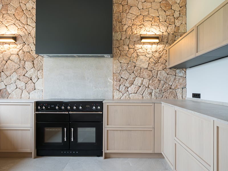 Cuisine contemporaine avec piano de cuisson Falcon noir, mur en pierre naturelle et meubles en bois clair.