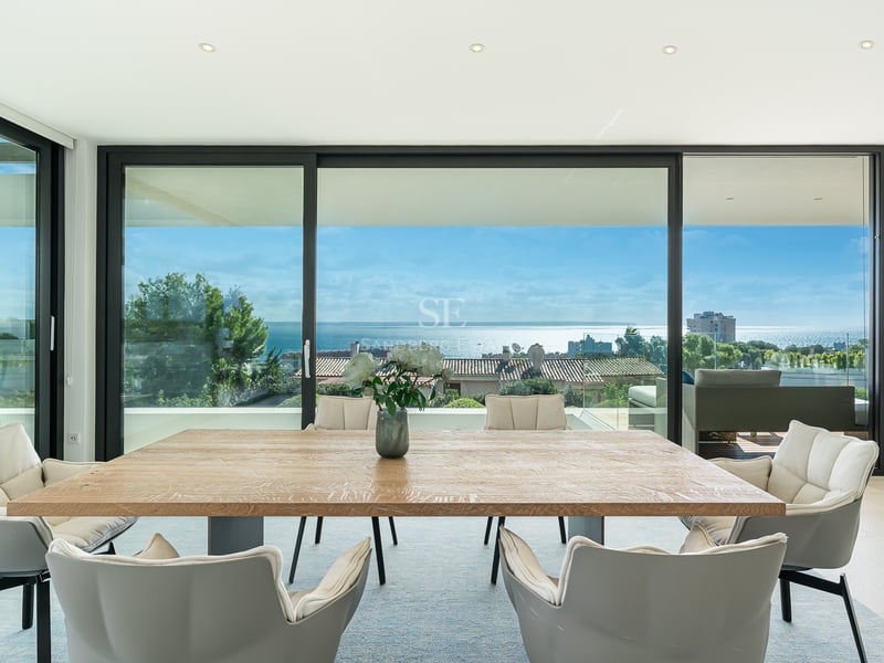 Salle à manger lumineuse avec table en bois et chaises grises face à des baies vitrées avec vue sur l'océan.