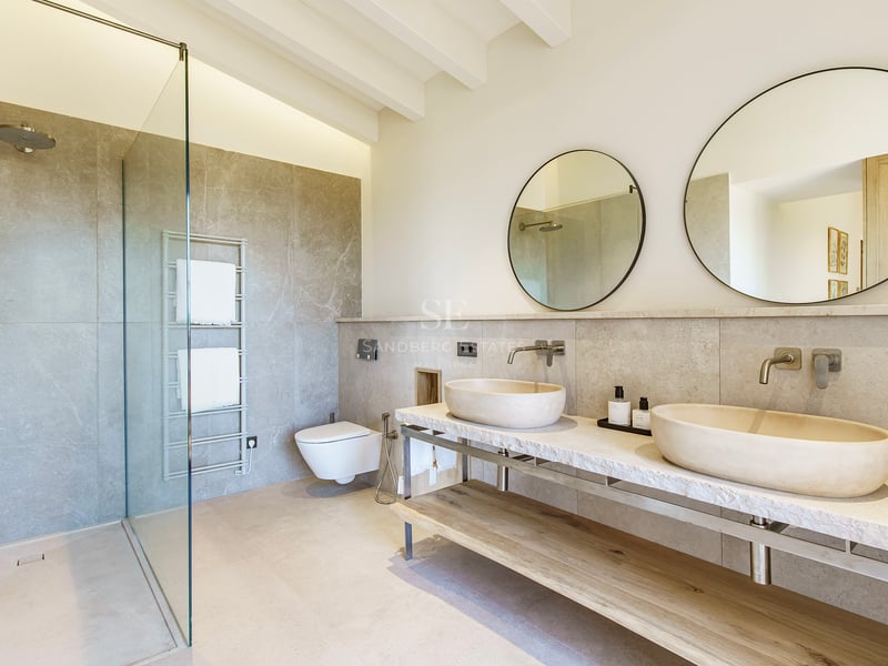Baño de lujo con dos lavabos de piedra sobre encimera de mármol, espejos circulares y ducha de cristal.