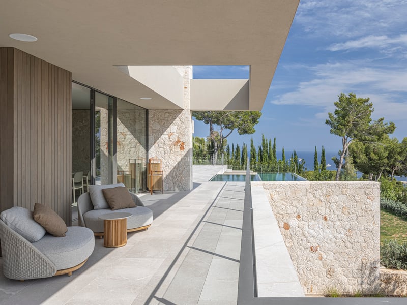 Terrasse de luxe avec fauteuils gris, murs en pierre et piscine à débordement surplombant la mer et les pins.