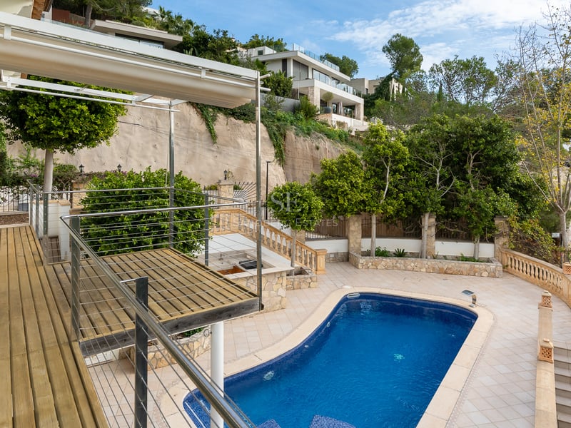 Vue plongeante sur une piscine bleue, une terrasse en bois et un patio carrelé entourés de verdure.