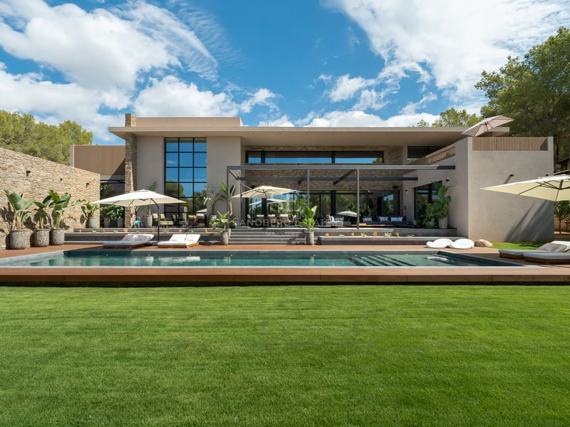 Villa contemporaine avec piscine allongée, terrasse en bois, pelouse verte et façade en pierre sous un ciel bleu.