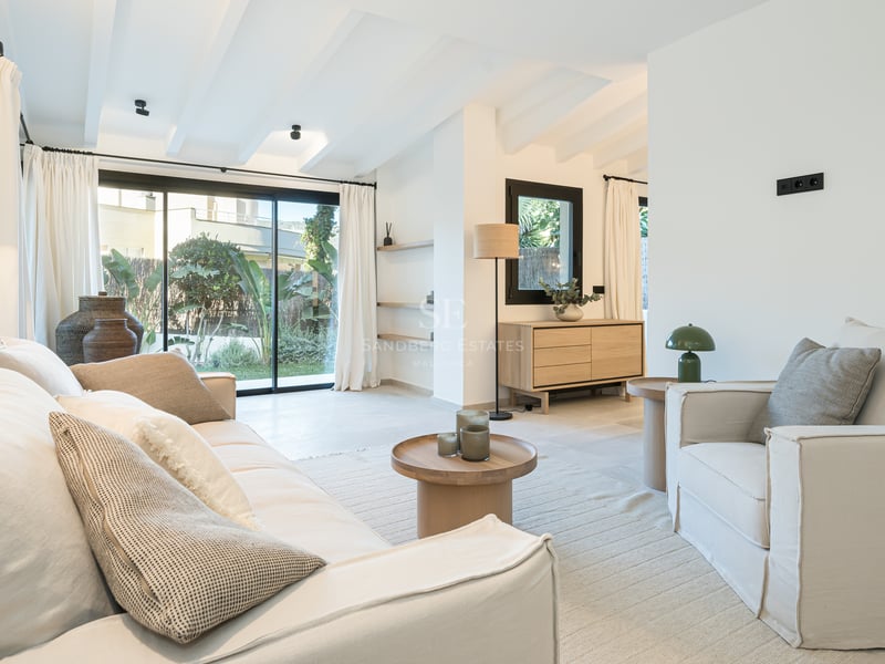 Salon lumineux avec plafonds à poutres blanches, canapés crème et baies vitrées ouvrant sur un jardin luxuriant.