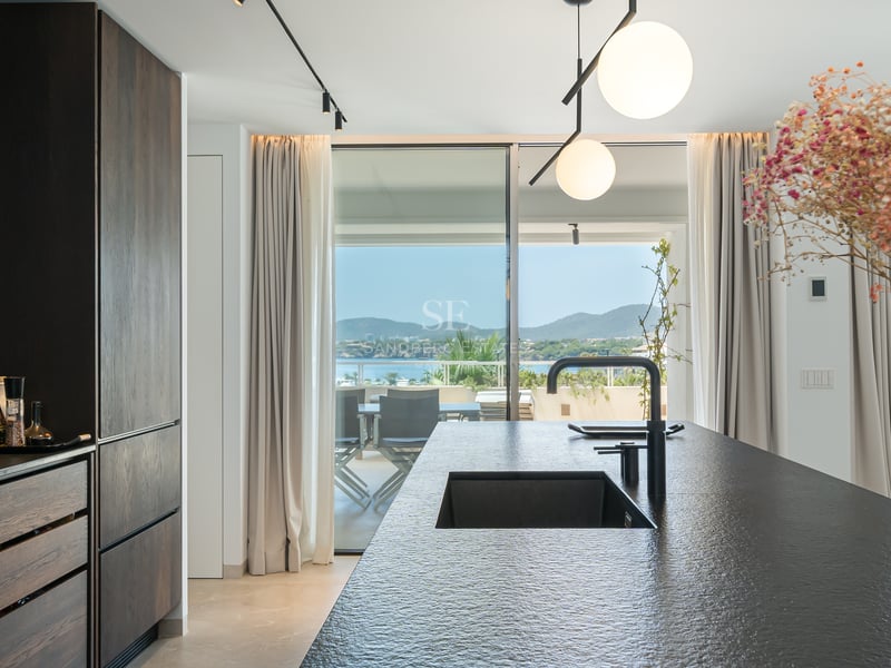 Cuisine moderne avec un îlot en pierre sombre et un évier noir donnant sur une terrasse ensoleillée et la mer.
