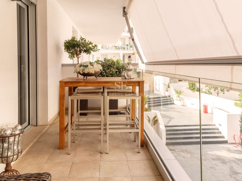 Terrasse moderne avec table haute en bois, tabourets, store blanc et garde-corps en verre donnant sur la cour.