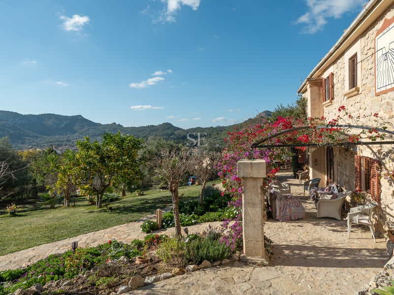 Terrasse en pierre avec mobilier extérieur, bougainvilliers roses et vue sur les montagnes sous un ciel bleu.