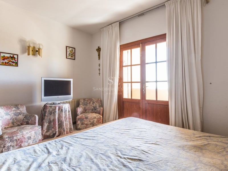 Chambre avec lit, deux fauteuils à fleurs et portes en bois ouvrant sur un balcon.