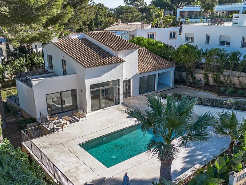 Vue aérienne d'une villa blanche avec toit en tuiles, piscine turquoise et grande terrasse entourée de pins et palmiers.