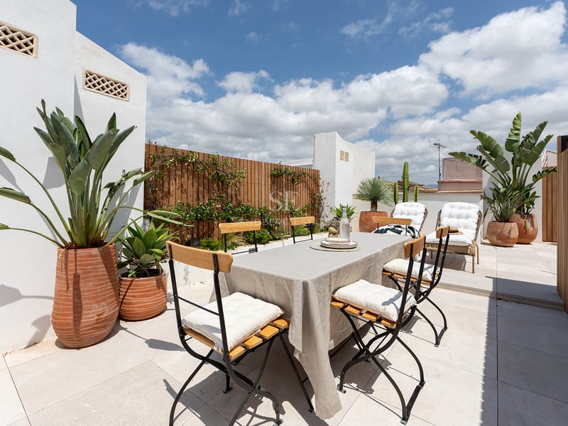 Terraza privada con mesa de comedor, mantelería de lino, valla de madera y plantas tropicales bajo cielo azul.