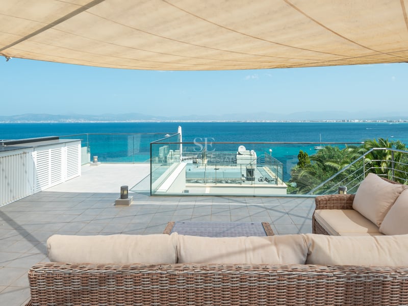 Toit-terrasse ensoleillé avec canapé en rotin, voile d'ombrage, garde-corps en verre et vue imprenable sur la mer.