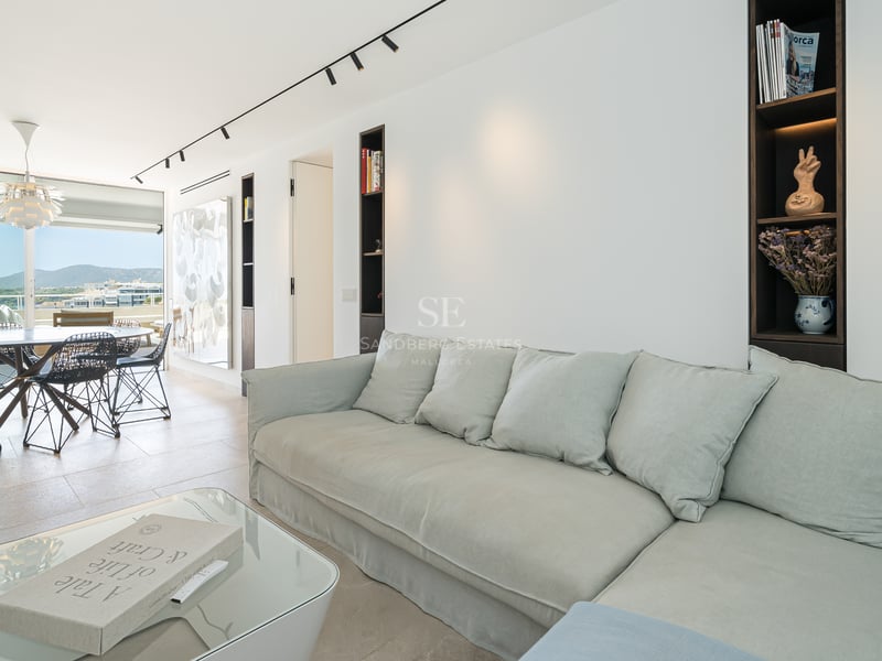 Salon lumineux avec un grand canapé gris clair, table basse en verre et salle à manger en arrière-plan avec vue sur la mer.