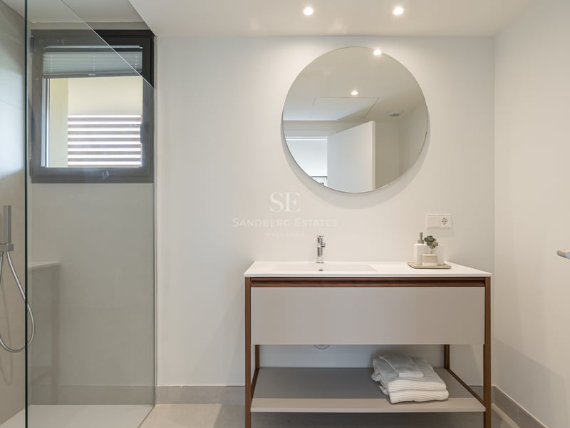 Baño de diseño minimalista con ducha de cristal, lavabo con mueble de madera y espejo redondo sobre pared blanca.