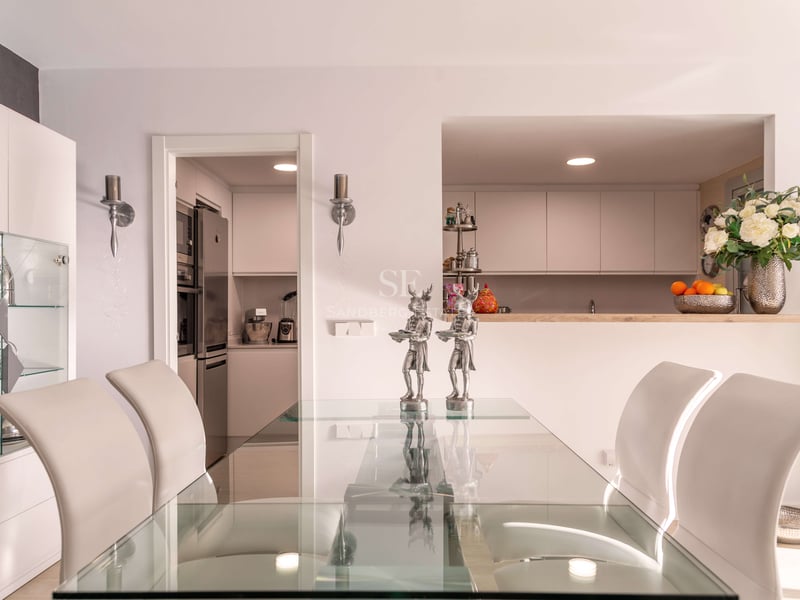 Salle à manger contemporaine avec table en verre, chaises blanches et vue sur une cuisine blanche moderne.