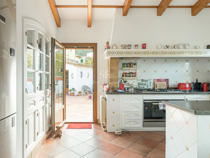 Cuisine avec poutres en bois, sol en terre cuite et porte ouverte sur une cour ensoleillée.