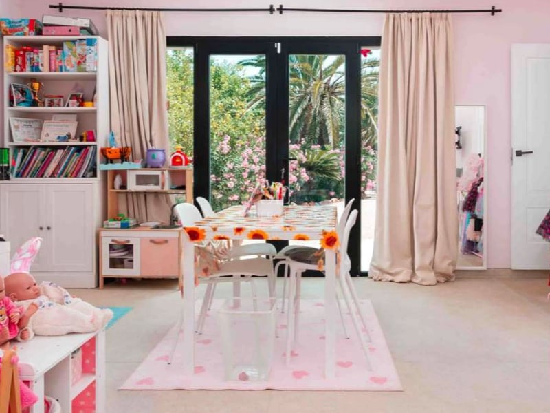 Une salle de jeux pour enfants aux murs roses, dotée de baies vitrées ouvrant sur un jardin avec palmiers.