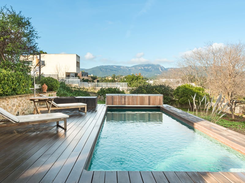 Piscine rectangulaire avec terrasse en bois sombre, bains de soleil blancs et vue panoramique sur les montagnes.