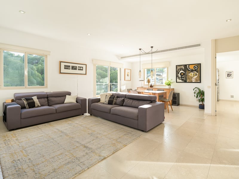 Grand salon lumineux avec deux canapés gris, carrelage clair et coin repas en arrière-plan avec de grandes fenêtres.
