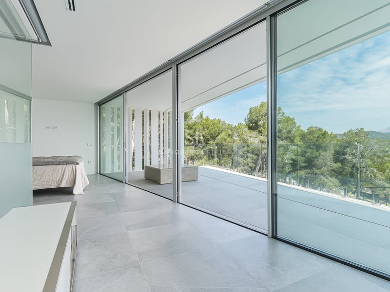 Chambre minimaliste avec de grandes baies vitrées coulissantes ouvrant sur une terrasse surplombant la forêt.