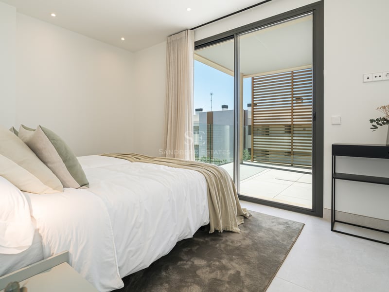 Dormitorio contemporáneo con cama blanca, consola negra y puertas correderas hacia una terraza soleada.