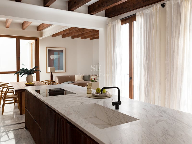 Cocina moderna con isla de mármol blanco, fregadero integrado, muebles de madera oscura y vigas de madera vistas.