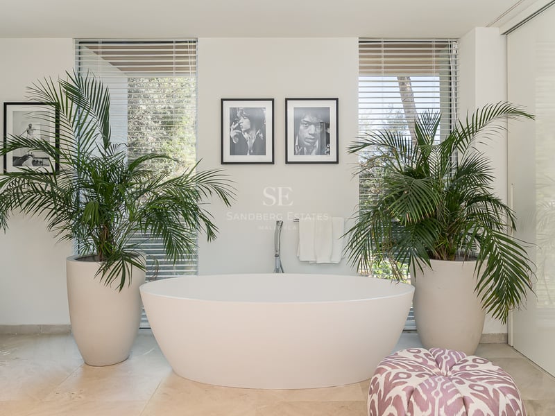 Baignoire îlot ovale blanche entourée de grands palmiers devant des fenêtres toute hauteur avec stores.
