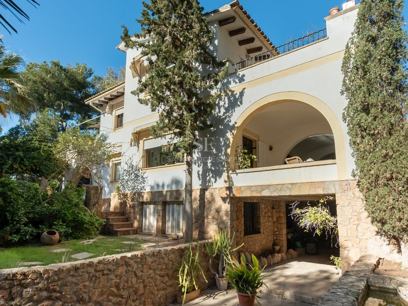 Villa méditerranéenne de trois étages avec balcon arqué, base en pierre naturelle et jardin mature sous un ciel bleu.