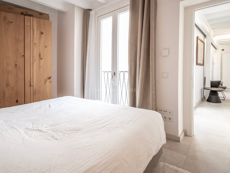 Chambre lumineuse avec lit blanc, grande armoire en bois et portes-fenêtres donnant sur un balcon.