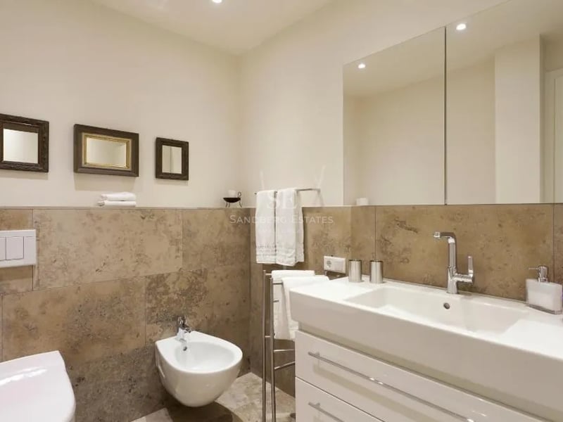 Salle de bain moderne avec carrelage en pierre beige, meuble vasque blanc, bidet et grand miroir mural.