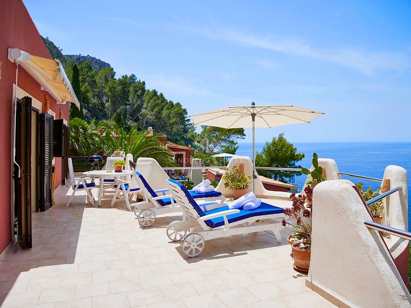 Terrasse ensoleillée avec chaises longues bleues, parasol blanc et vue imprenable sur la mer et les falaises.