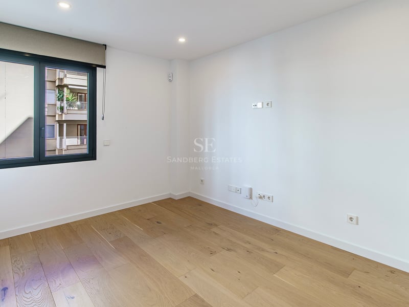 Habitación vacía y luminosa con suelos de madera clara, paredes blancas y un gran ventanal con persiana.