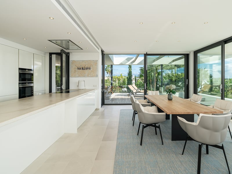 Salle à manger moderne avec table en bois, chaises grises et cuisine blanche, ouverte sur une terrasse par des baies vitrées.