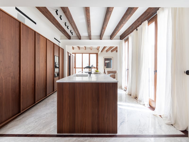 Cocina de lujo con isla de madera alistonada, encimeras de mármol y vigas de madera oscura bajo luz natural.