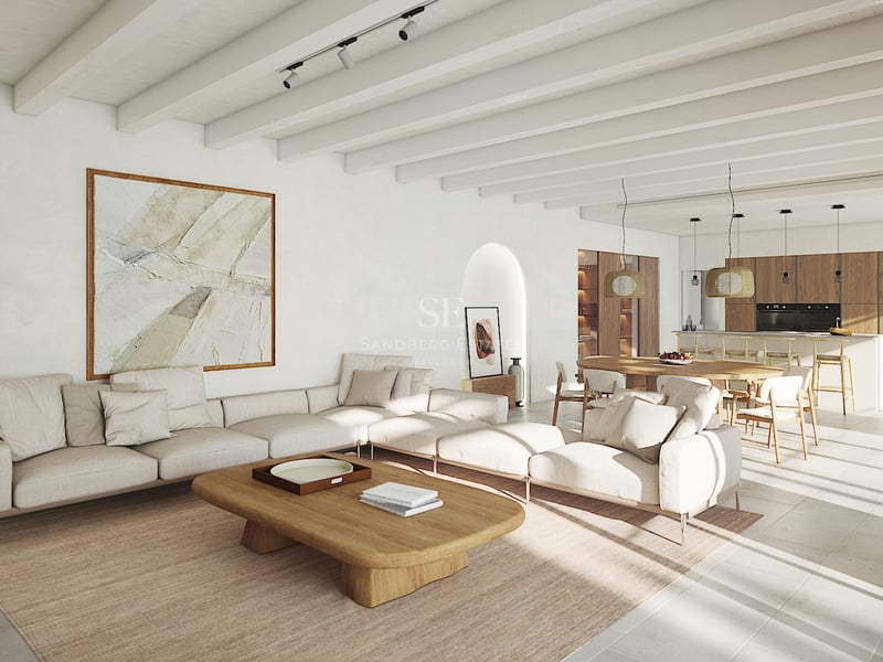 Salón luminoso de planta abierta con vigas blancas, sofá modular, mesa de madera y cocina integrada al fondo.