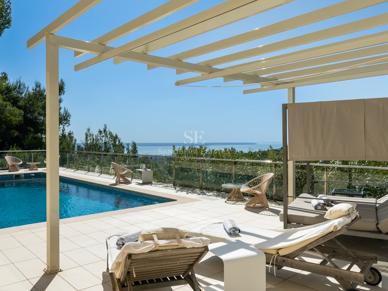 Terrasse ensoleillée avec piscine, pergola blanche, transats et garde-corps en verre surplombant la mer.