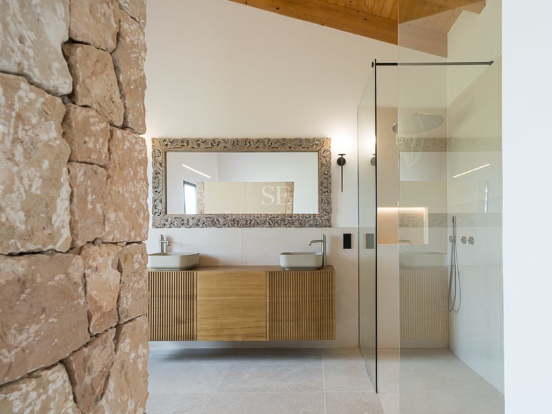 Salle de bains moderne avec mur en pierre brute, plafond en bois, douche vitrée et double vasque suspendue.