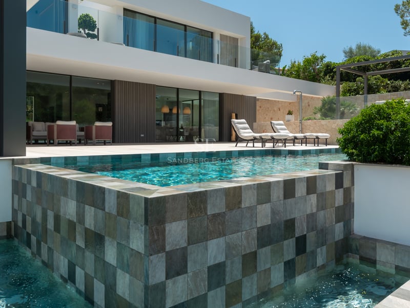Villa blanche moderne avec piscine à débordement en pierre, chaises longues et grandes baies vitrées.