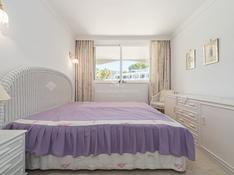 Chambre avec tête de lit en osier blanc, couvre-lit violet et fenêtre offrant une vue sur l'extérieur.