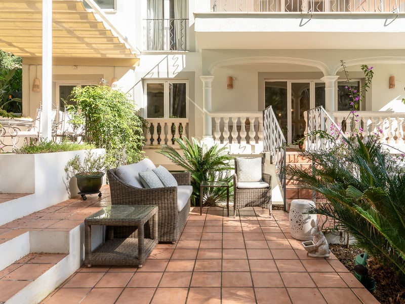 Terrasse ensoleillée en terre cuite avec mobilier en osier et plantes devant une façade de villa blanche.