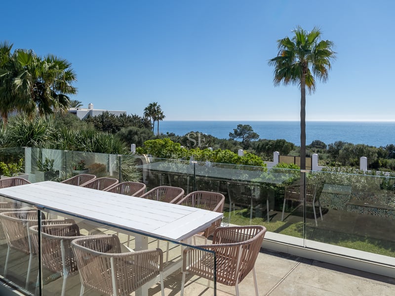 Grande terrasse avec table blanche, chaises design et garde-corps en verre surplombant la mer Méditerranée et des palmiers.