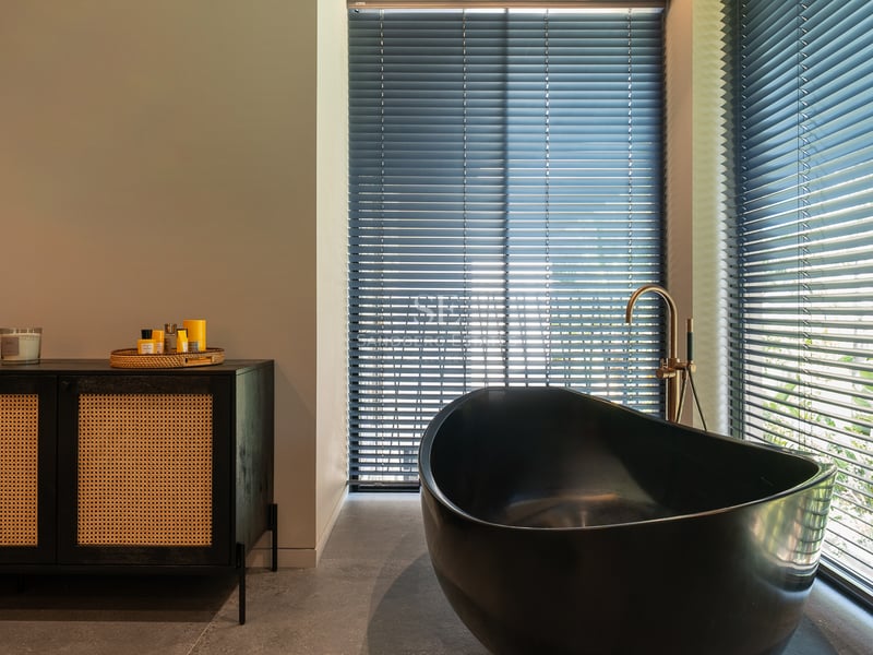 Salle de bain de luxe avec baignoire ovale noire, robinetterie dorée et buffet en rotin près de fenêtres à stores.